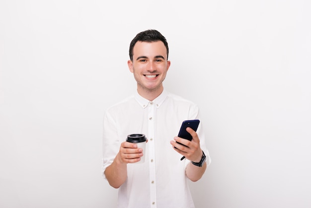 Jovem alegre está olhando para a câmera segurando uma xícara com bebida quente e um telefone em um fundo branco.