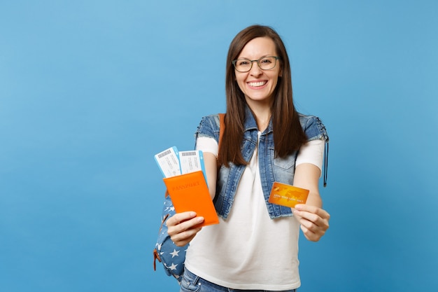 Jovem alegre feliz estudante de óculos com mochila segurar o cartão de crédito do bilhete de embarque do passaporte isolado sobre fundo azul. educação em faculdade universitária no exterior. conceito de voo de viagens aéreas.