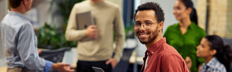 Jovem alegre homem de raça mista segurando tablet digital e sorrindo para a câmera enquanto trabalha com colegas no escritório moderno foco seletivo Pessoas de negócios no trabalho