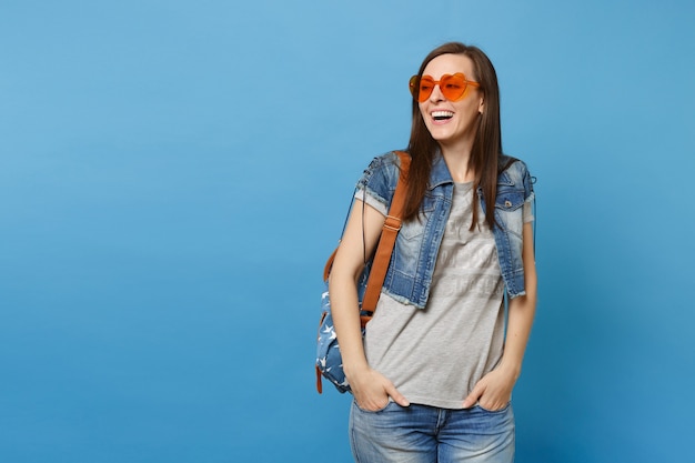 Jovem alegre linda estudante com mochila usando óculos em forma de coração laranja, olhando de lado, de mãos dadas nos bolsos isolados sobre fundo azul. Educação no ensino médio. Copie o espaço para anúncio.