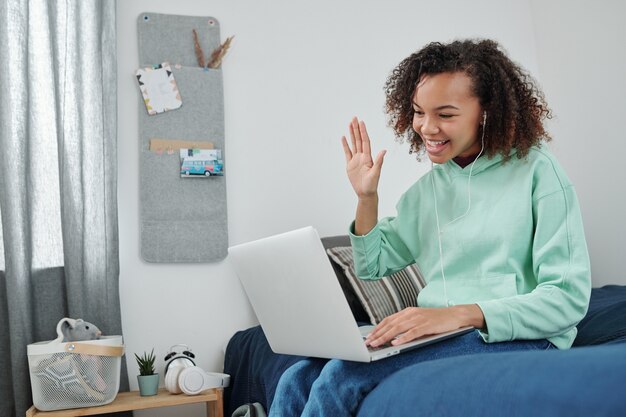 Jovem alegre mestiça em trajes casula acenando com a mão para a amiga na tela do laptop enquanto se comunica por vídeo chat em casa