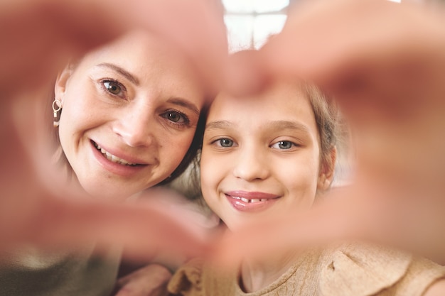 Jovem alegre morena e sua linda filha olhando para você com sorrisos através do coração de mão em pé na frente da câmera