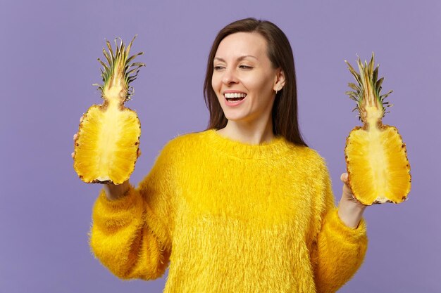 Jovem alegre na camisola de pele olhando de lado segurando metades de frutas frescas de abacaxi maduro isoladas em fundo pastel violeta. Estilo de vida vívido de pessoas, relaxe o conceito de férias. Mock up espaço de cópia.