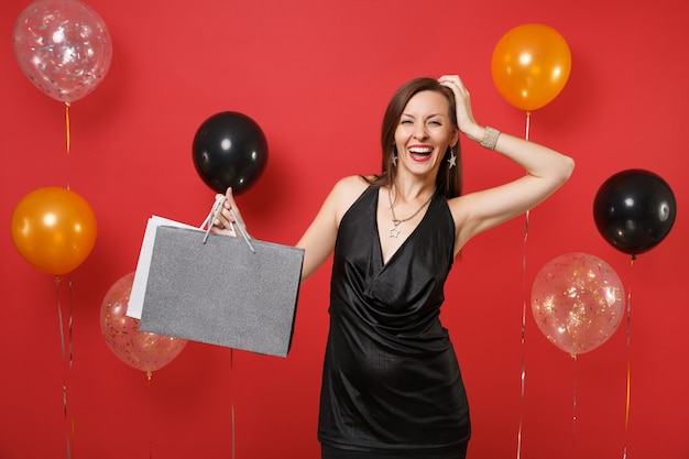 Jovem alegre num vestido preto, colocando a mão na cabeça, segurando os pacotes com as compras depois de fazer compras em balões de ar de fundo vermelho brilhante. Feliz Ano Novo, conceito de festa de feriado de maquete de aniversário.