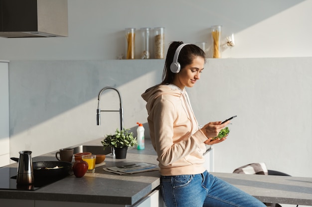 Jovem alegre ouvindo música com fones de ouvido na cozinha em casa, comendo salada em uma tigela