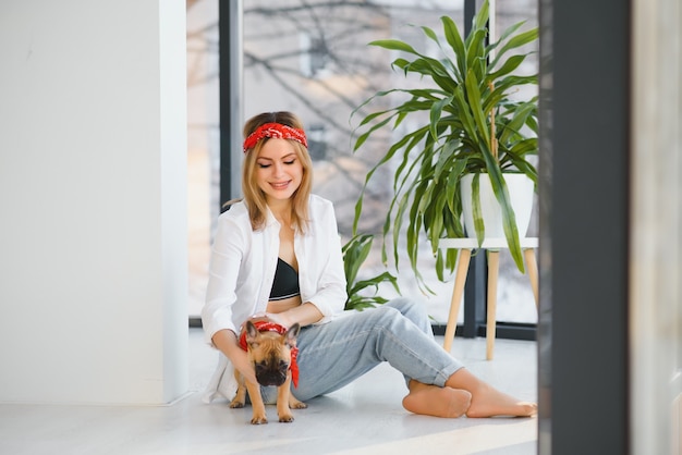 Jovem alegre segurando seu cachorrinho com nariz preto e rindo. Retrato interno de menina sorridente posando com buldogue francês