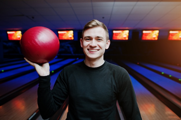 Jovem alegre segurando uma bola de boliche e sorrindo para a câmera em pé contra pistas de boliche com luz ultravioleta.