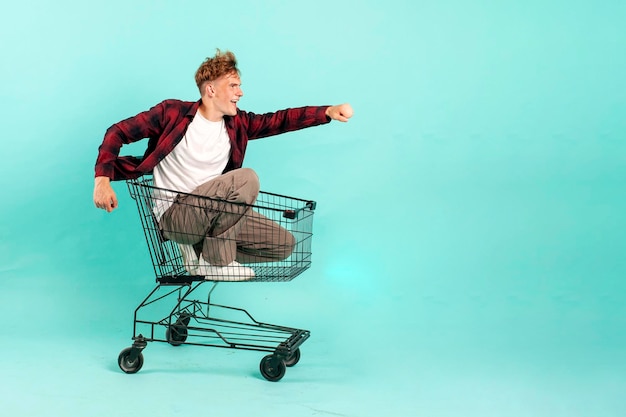 Foto jovem alegre senta-se no carrinho de compras e mostra um gesto para a frente em fundo azul