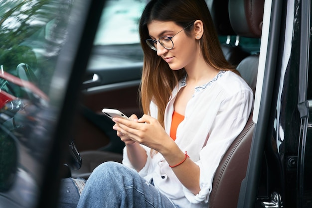 Jovem alegre sentada no carro no banco do motorista e olhando para a tela do celular