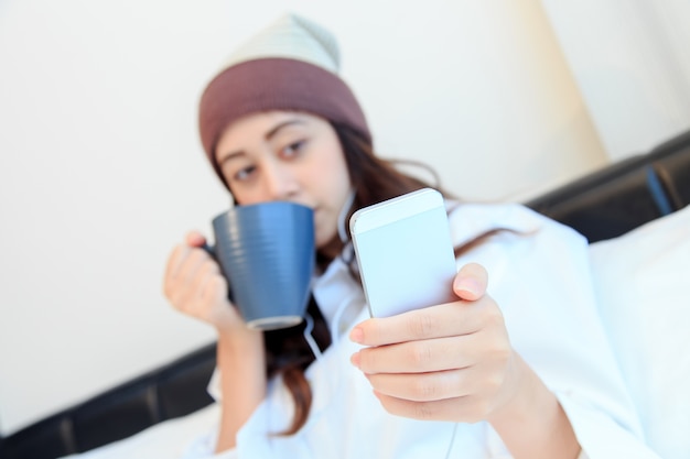 Jovem alegre sentado na cama mensagens de texto em seu smartphone e beber café em casa no quarto