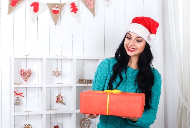 Jovem alegre sentado no interior de ano novo vestido com chapéu de Papai Noel segurando a caixa de presente vermelha.