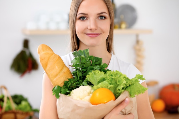 Jovem alegre sorridente está pronta para cozinhar em uma cozinha Dona de casa está segurando um grande saco de papel cheio de frutas e legumes frescos e olhando para a câmera