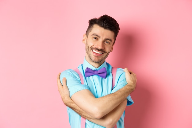 Jovem alegre, sorrindo e se abraçando, enrole as mãos ao redor do próprio corpo e pareça feliz, em pé sobre um fundo rosa.