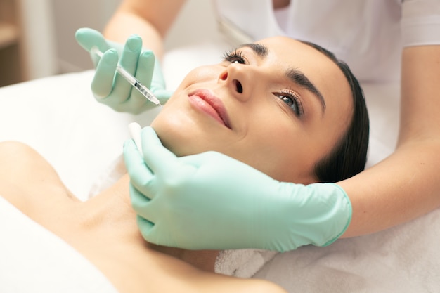 Jovem alegre sorrindo enquanto cosmetologista profissional injetando na pele do rosto