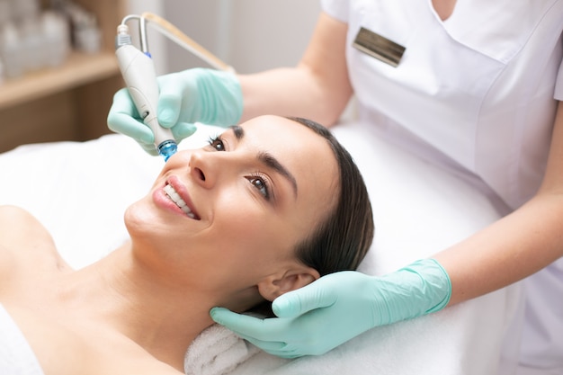 Jovem alegre sorrindo enquanto cosmetologista profissional limpando os poros da pele do rosto com a ajuda de uma ferramenta moderna