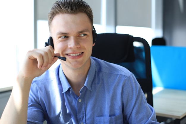 Jovem alegre suporte operador masculino de telefone no fone de ouvido, no local de trabalho, enquanto estiver usando o laptop, serviço de ajuda e conceito de centro de chamada de consultoria de cliente.