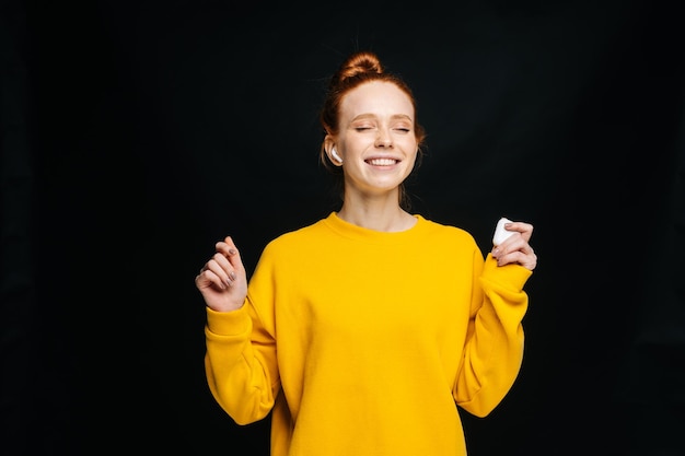 Jovem alegre usando fones de ouvido sem fio dançando enquanto ouve música popular em fundo preto isolado olhos fechados Modelo de senhora ruiva bonita mostrando emocionalmente expressões faciais