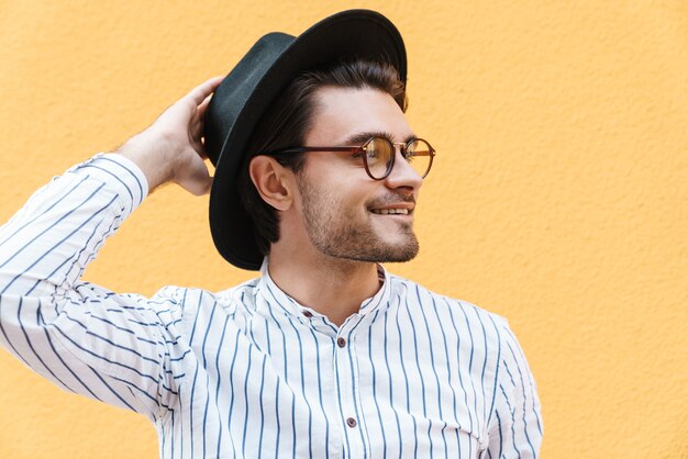 jovem alegre usando óculos e chapéu preto, sorrindo e olhando de lado para copyspace isolado na parede amarela