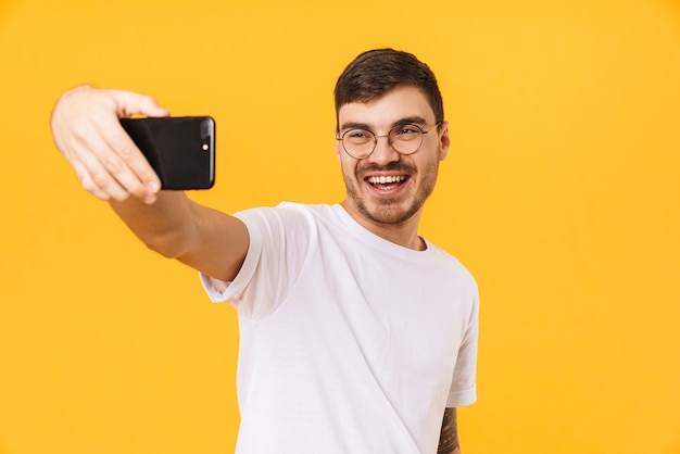 jovem alegre usando óculos tirando selfie no celular e sorrindo isolado na parede amarela