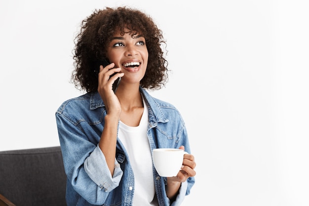 Foto jovem alegre, vestindo roupas jeans casuais, sentada em uma cadeira isolada na parede branca, segurando uma xícara de café, usando um telefone celular