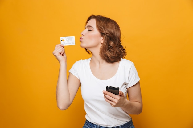 Foto jovem alegre vestindo uma camiseta em pé, isolada na parede amarela, usando um telefone celular, mostrando um cartão de crédito de plástico