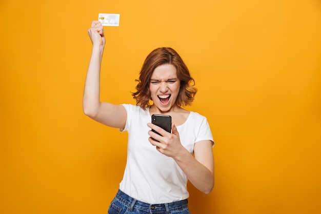 Jovem alegre vestindo uma camiseta em pé, isolada na parede amarela, usando um telefone celular, mostrando um cartão de crédito de plástico