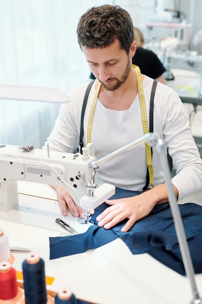 Jovem alfaiate sério se concentrando no trabalho de costura enquanto está sentado diante da máquina na oficina e terminando com o pedido