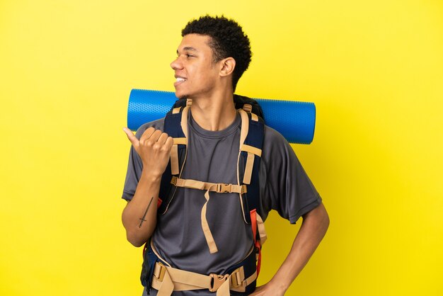 Jovem alpinista afro-americano com uma grande mochila isolada em um fundo amarelo apontando para o lado para apresentar um produto