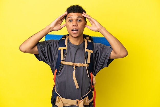 Jovem alpinista afro-americano com uma grande mochila isolada em um fundo amarelo com expressão de surpresa