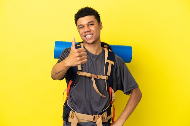 Jovem alpinista afro-americano com uma grande mochila isolada em um fundo amarelo com polegares para cima porque algo bom aconteceu