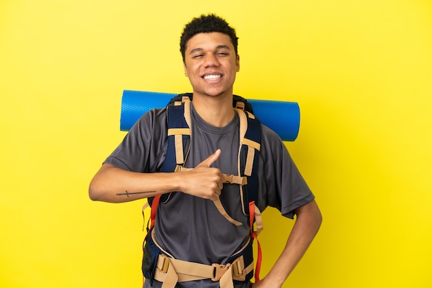 Jovem alpinista afro-americano com uma grande mochila isolada em um fundo amarelo e fazendo um gesto de polegar para cima