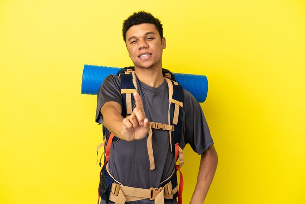 Jovem alpinista afro-americano com uma grande mochila isolada em um fundo amarelo, mostrando e levantando um dedo