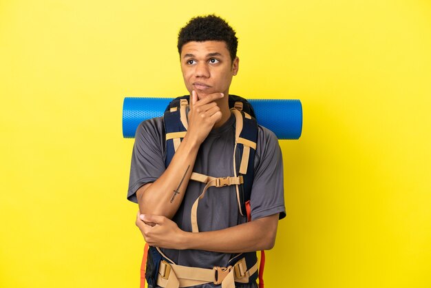 Jovem alpinista afro-americano com uma grande mochila isolada em um fundo amarelo, tendo dúvidas e pensando