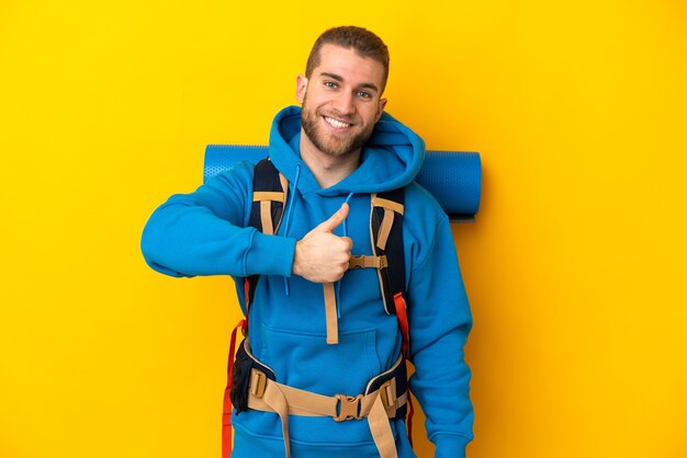 Jovem alpinista caucasiano com uma grande mochila isolada em amarelo fazendo um gesto de polegar para cima