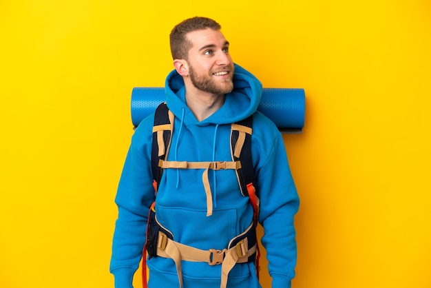 Jovem alpinista caucasiano com uma grande mochila isolada em amarelo pensando uma ideia enquanto olha para cima