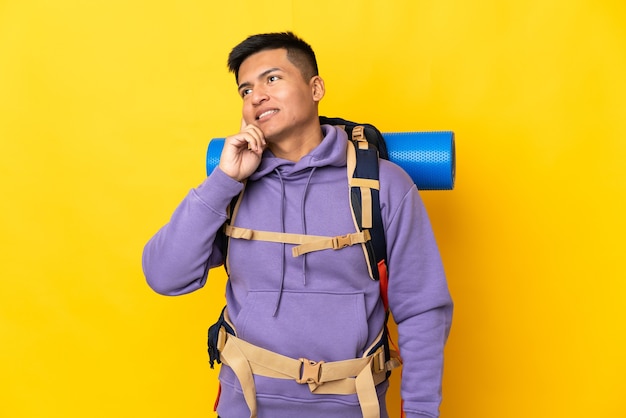 Jovem alpinista com uma grande mochila isolada em um fundo amarelo pensando uma ideia enquanto olha para cima