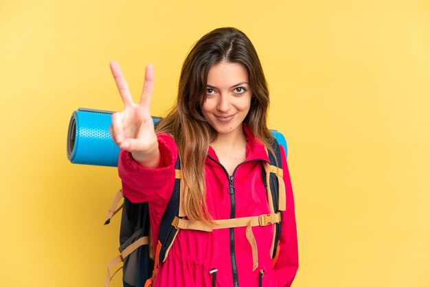 Jovem alpinista com uma grande mochila isolada em um fundo amarelo sorrindo e mostrando sinal de vitória