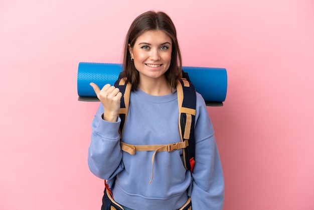 Jovem alpinista com uma grande mochila isolada em um fundo rosa apontando para o lado para apresentar um produto