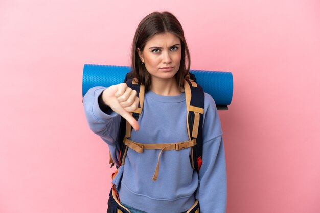 Jovem alpinista com uma grande mochila isolada em um fundo rosa, mostrando o polegar para baixo com expressão negativa