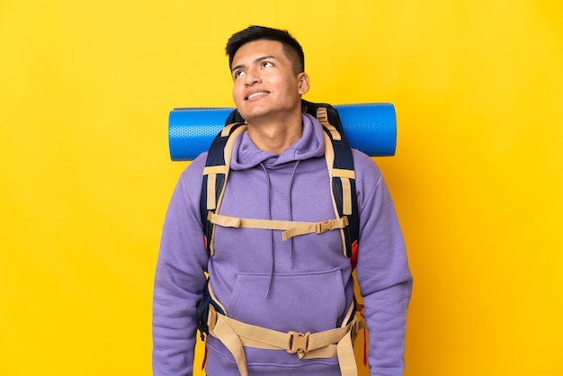 Jovem alpinista com uma grande mochila isolada na parede amarela pensando uma ideia enquanto olha para cima