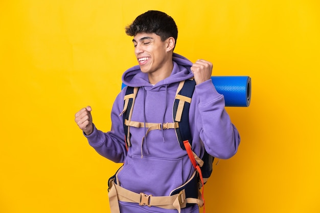 Jovem alpinista com uma grande mochila sobre um fundo amarelo isolado, comemorando uma vitória