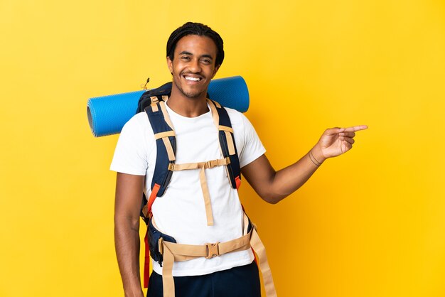 Jovem alpinista de tranças com uma grande mochila isolada em fundo amarelo apontando o dedo para o lado e apresentando um produto