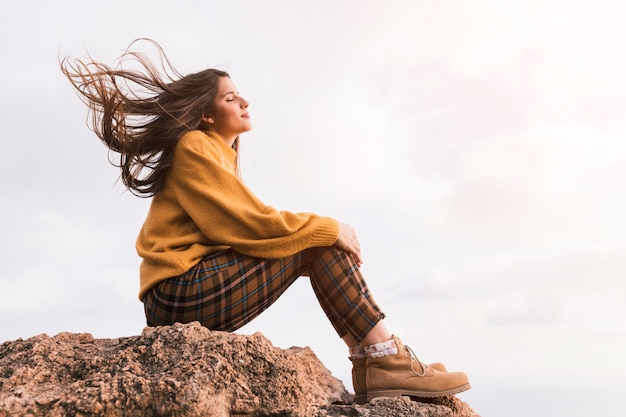 Jovem alpinista feminina sentada em cima de rocha, apreciando o ar fresco contra o céu