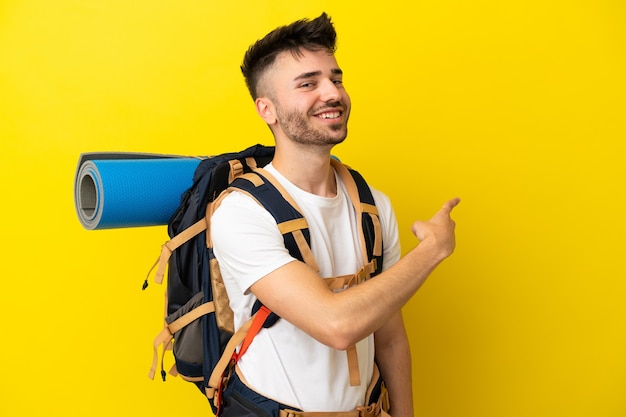 Jovem alpinista homem caucasiano com uma grande mochila isolada em um fundo amarelo apontando para trás