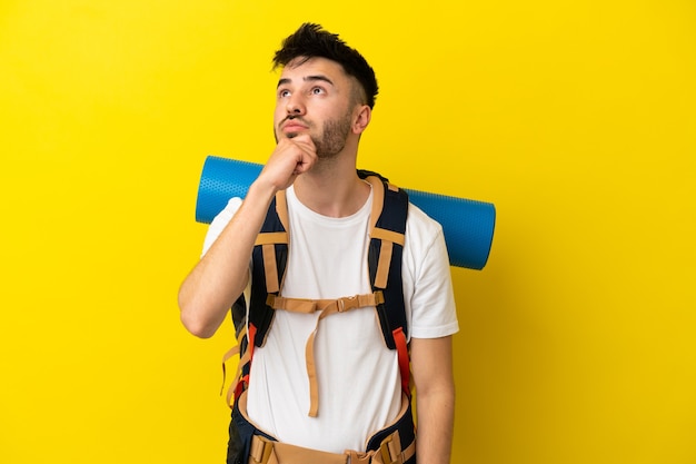 Jovem alpinista homem caucasiano com uma grande mochila isolada em um fundo amarelo e olhando para cima