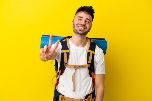 Jovem alpinista homem caucasiano com uma grande mochila isolada em um fundo amarelo sorrindo e mostrando sinal de vitória