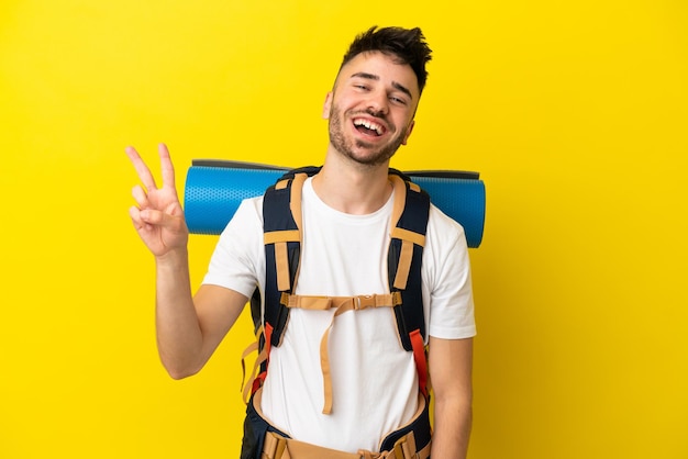 Jovem alpinista homem caucasiano com uma grande mochila isolada em um fundo amarelo sorrindo e mostrando sinal de vitória