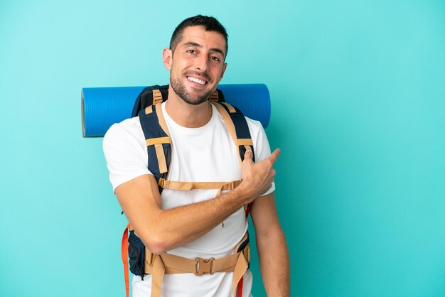 Jovem alpinista homem caucasiano com uma grande mochila isolada em um fundo azul apontando para trás