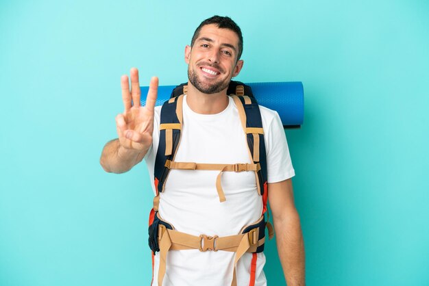 Jovem alpinista homem caucasiano com uma grande mochila isolada em um fundo azul feliz e contando três com os dedos