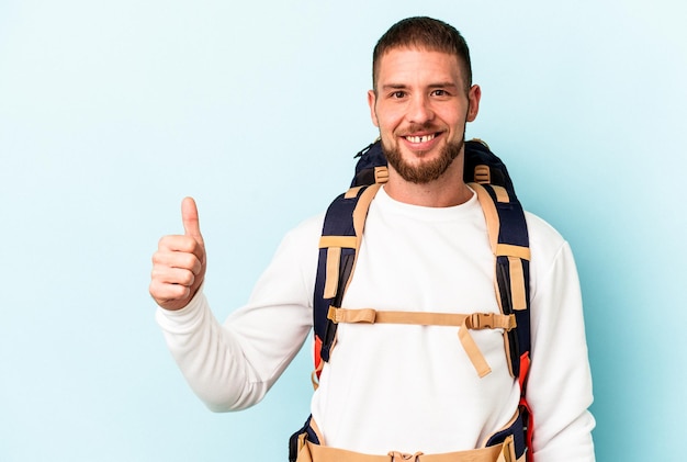 Jovem alpinista homem caucasiano isolado em um fundo azul, sorrindo e levantando o polegar